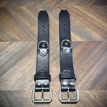 Top-down flat lay of a pair of unbuckled 1.5" black leather straps showcasing symmetric adjustment holes, centered steel D-rings, and roller buckles.