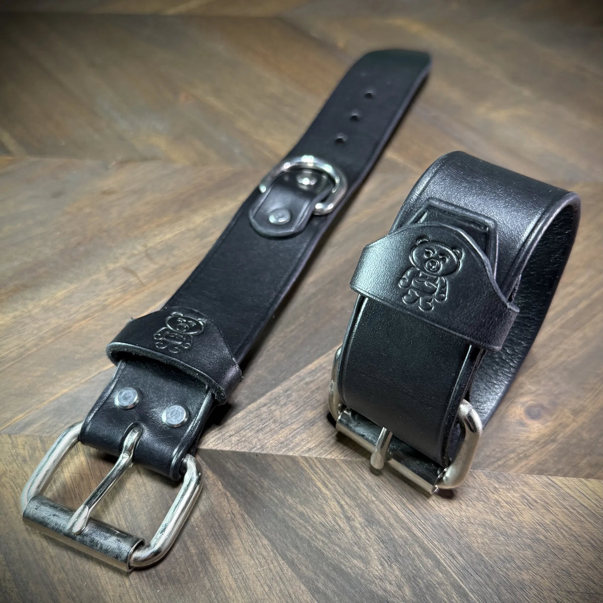 Hero tilt of a pair of handcrafted 1.5" black leather cuffs with one laid flat and one buckled, highlighting the steel hardware and premium veg-tan hide.