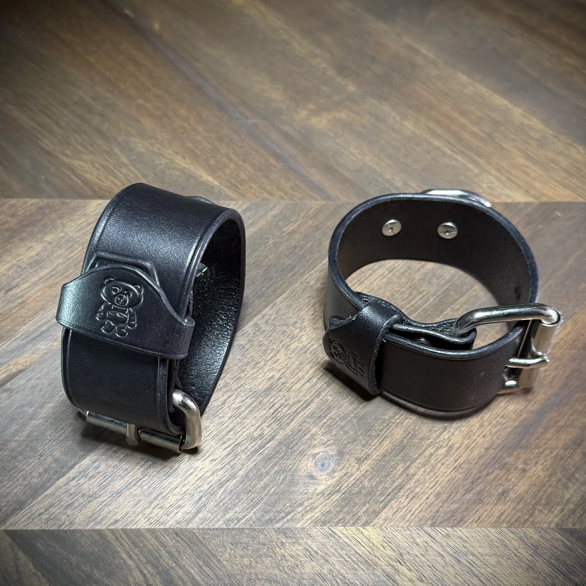 Detail view of two buckled 1.5" black leather wrist cuffs focusing on the debossed bear logo keeper loop and interior steel securing rivets.