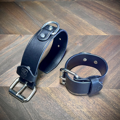 Three-quarter perspective of a pair of 1.5" black leather wrist restraints demonstrating leather thickness and structural steel buckle closure.