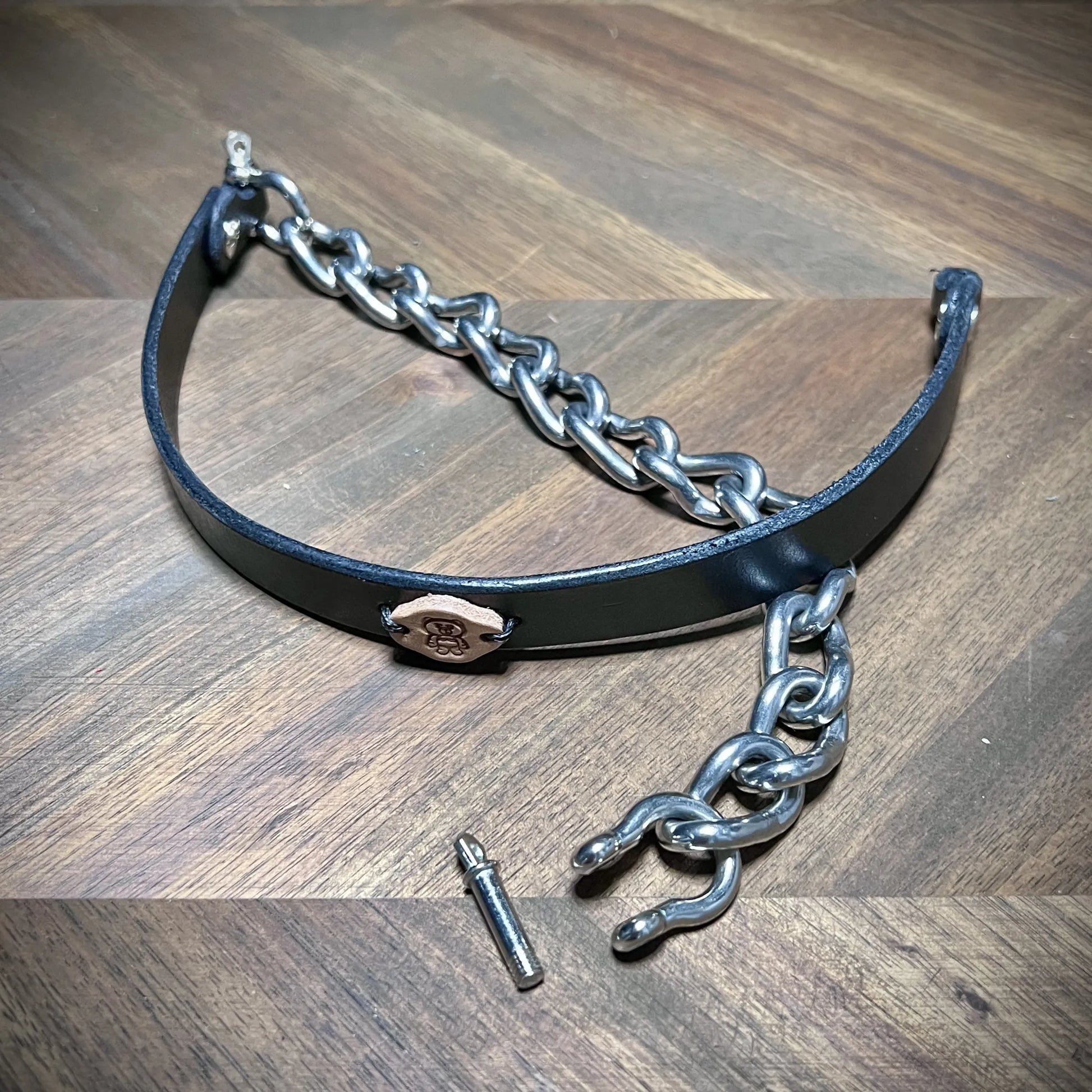 Macro view of an uncoupled black leather choker collar showing the removable screw-pin shackle closure mechanism in nickel-plated steel.