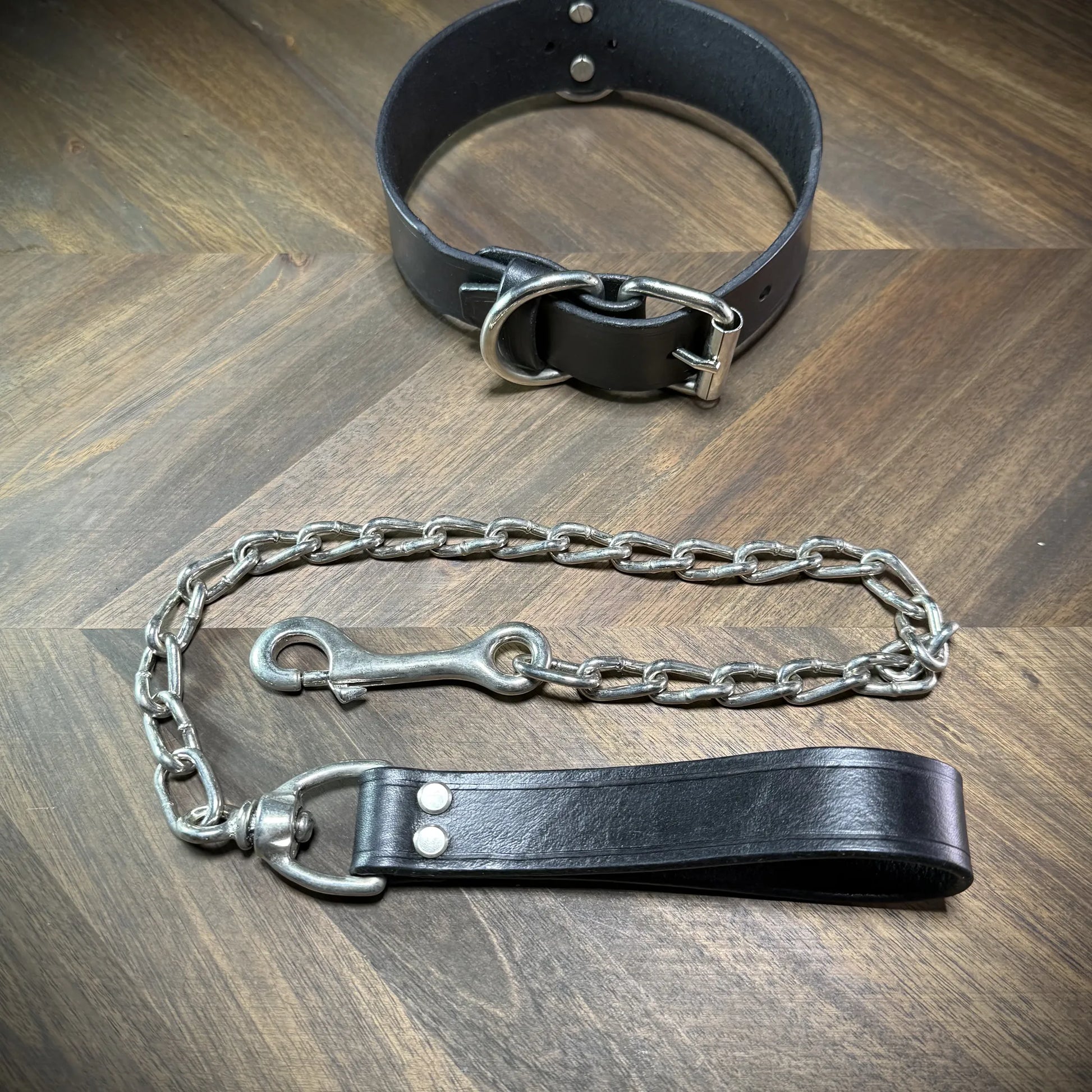 Hero-tilt view of a handcrafted 1.75" black leather collar with nickel-plated steel hardware and an attached heavy chain leash.