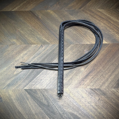 Studio-craft view of a Knotty Oso impact martinet handcrafted from black leather, featuring a hand-wrapped handle and multiple heavy straps.