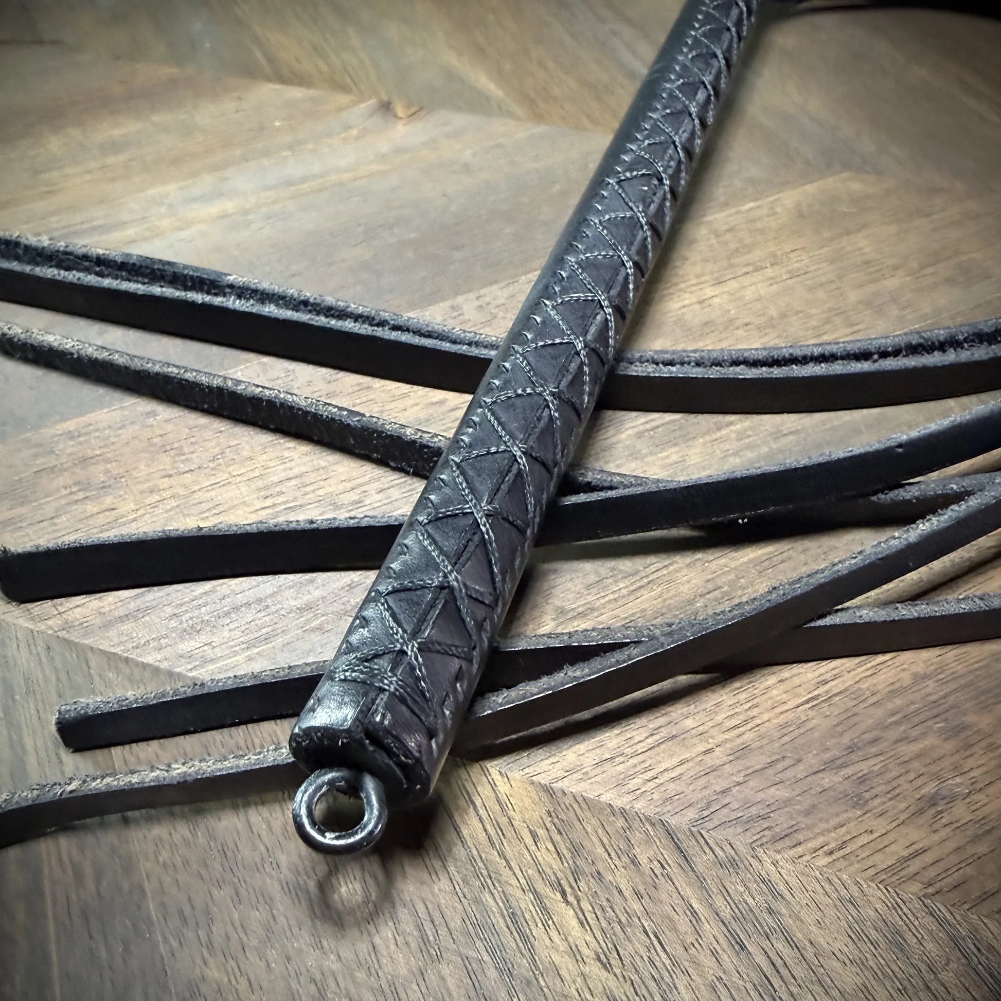 Detail-macro view of a black leather handle with a precision hand-sewn cross-stitch pattern, highlighting the integrated steel pommel ring.