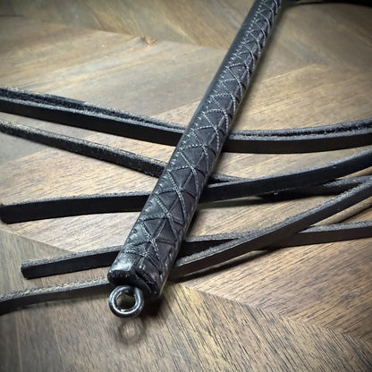Detail-macro view of a black leather handle with a precision hand-sewn cross-stitch pattern, highlighting the integrated steel pommel ring.