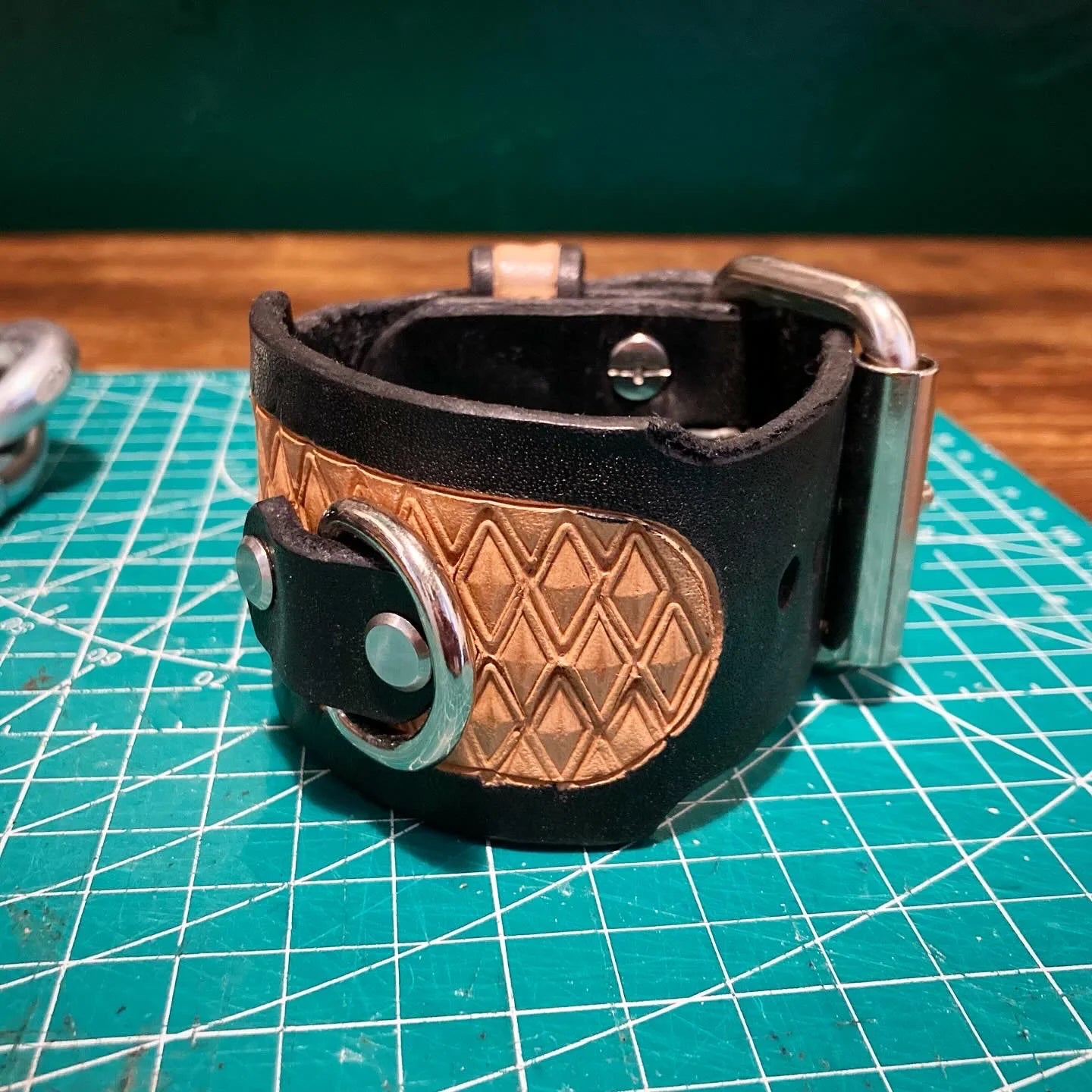 Hand-stamped geometric diamond leather cuff overlay with integrated heavy steel roller buckle on a green mat.