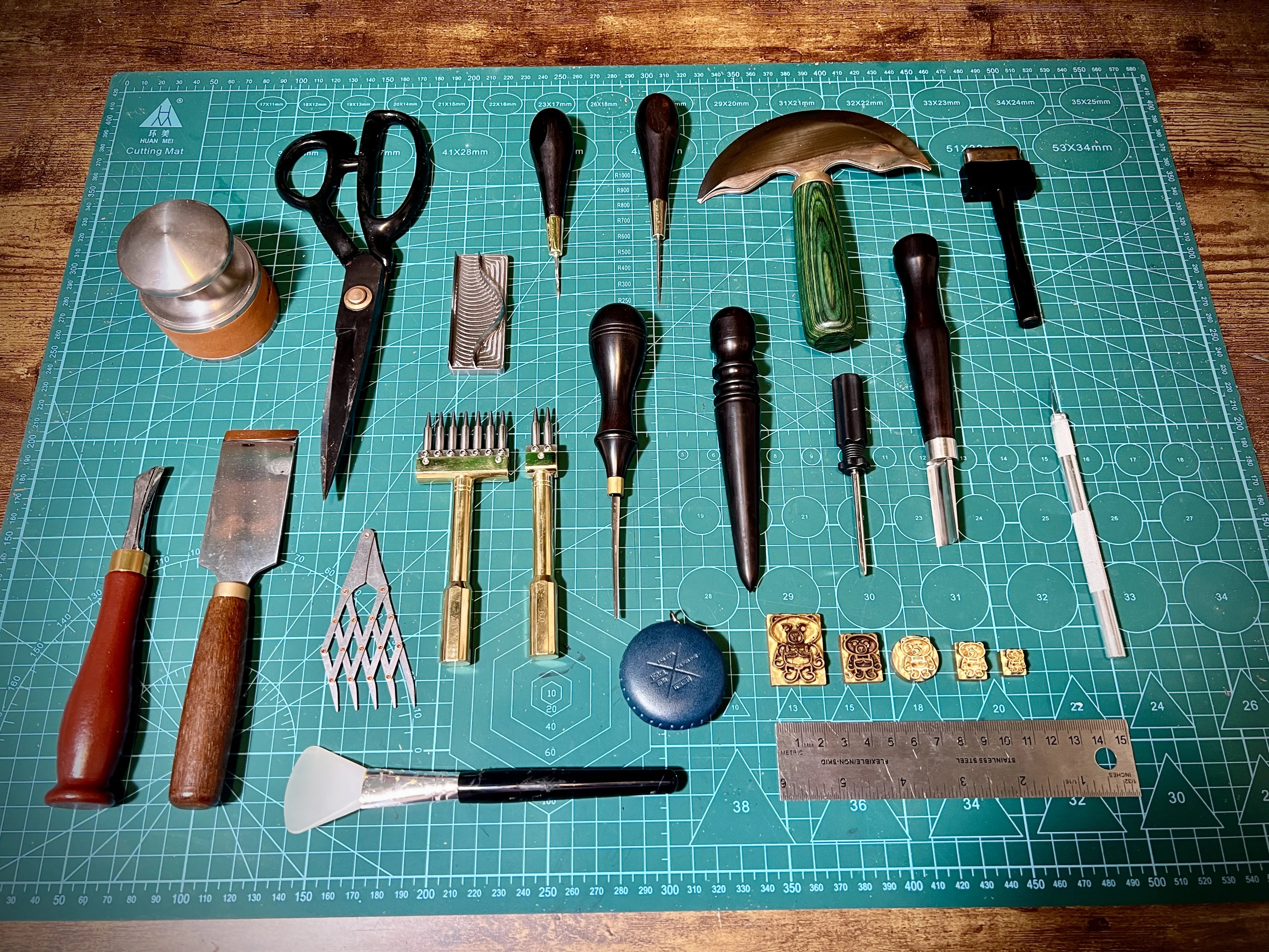 Symmetrical overhead layout of artisan leatherworking hand tools including a round knife, strap cutters, bevelers, and customized bear maker stamps on a cutting grid.