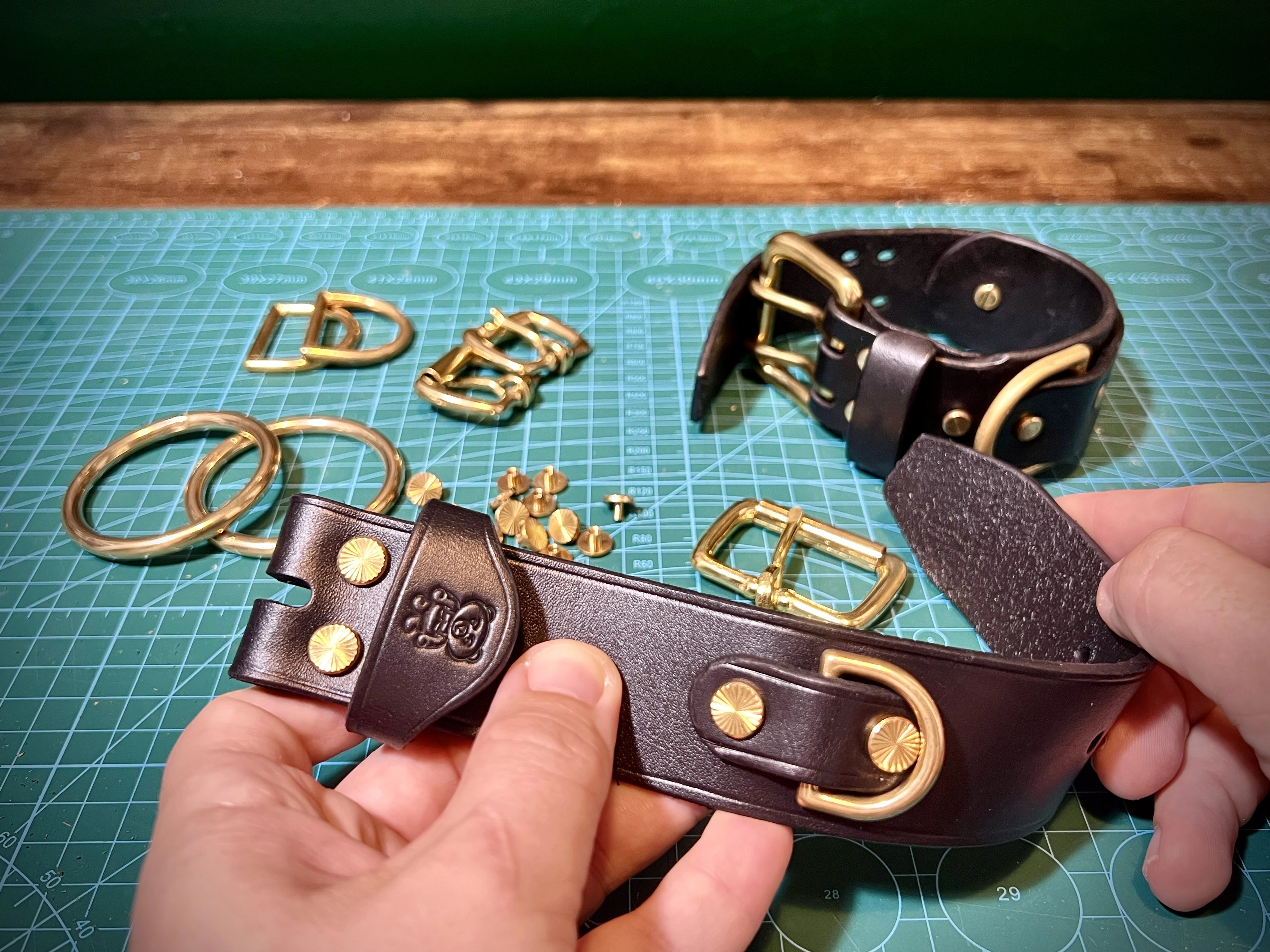 Artisan hands displaying a handcrafted black leather cuff with a stamped bear logo alongside solid brass hardware, buckles, and rivets on a green workshop cutting mat.