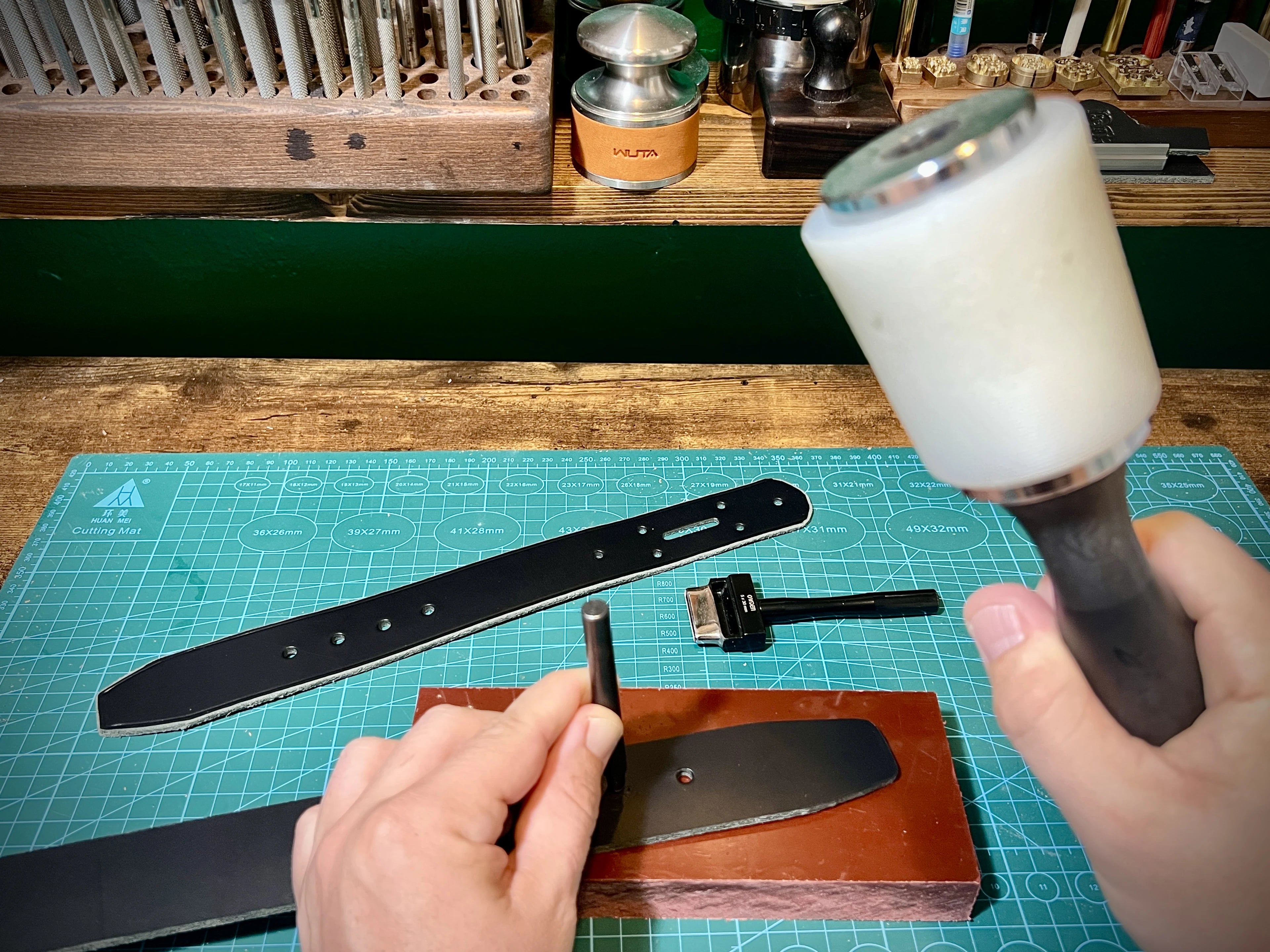 Artisan hands precisely aligning a steel hole drive punch over a premium black leather strap on a green grid mat next to a heavy white malleting maul.