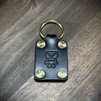 Vertical view of a handcrafted black leather keychain with solid brass hardware, featuring an embossed bear logo and starburst accent screws.