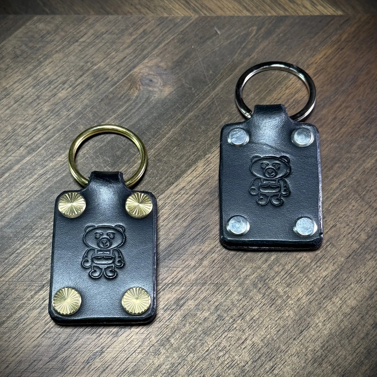 Studio craft view showcasing two black leather keychains side by side to compare the solid brass and nickel-plated steel hardware options.