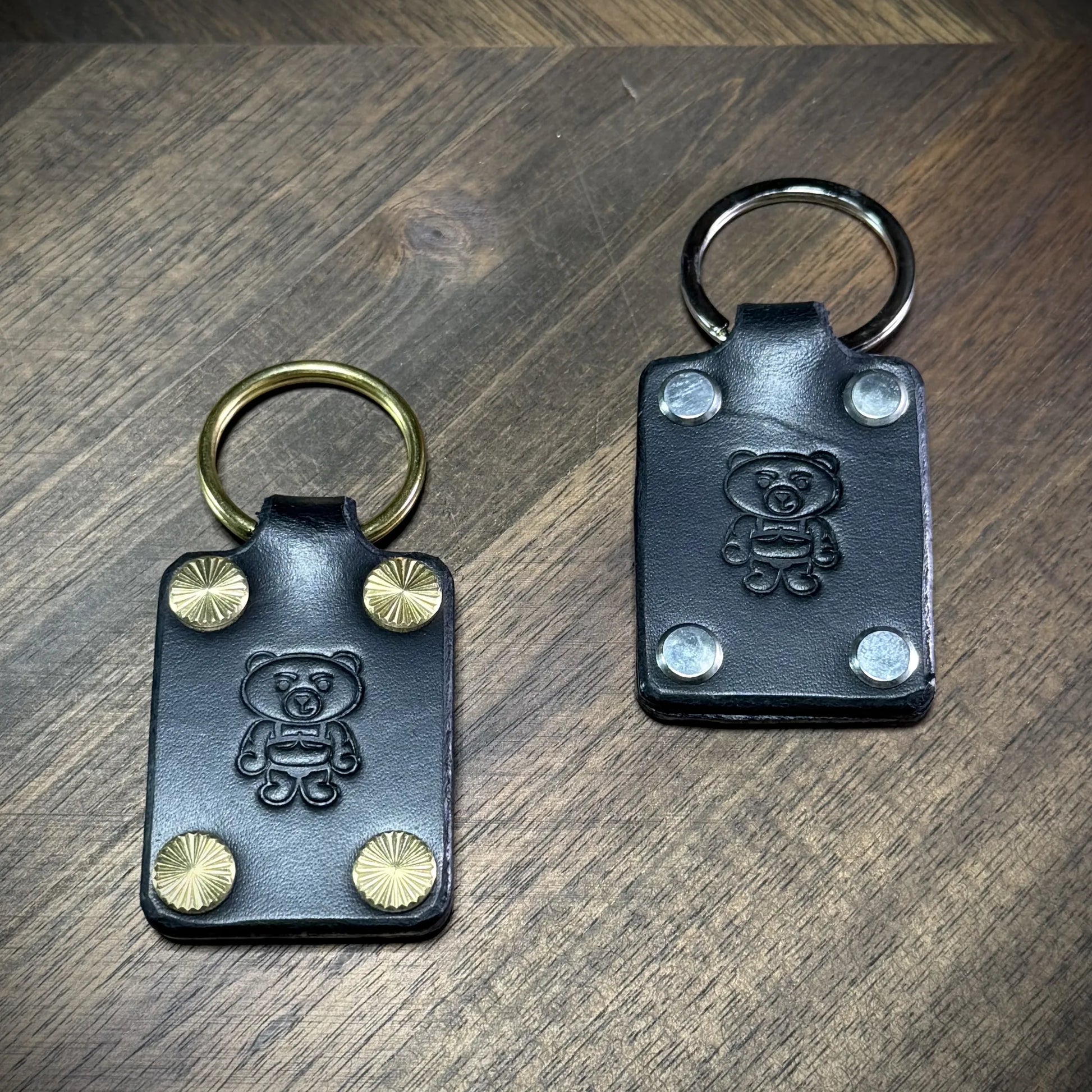 Studio craft view showcasing two black leather keychains side by side to compare the solid brass and nickel-plated steel hardware options.