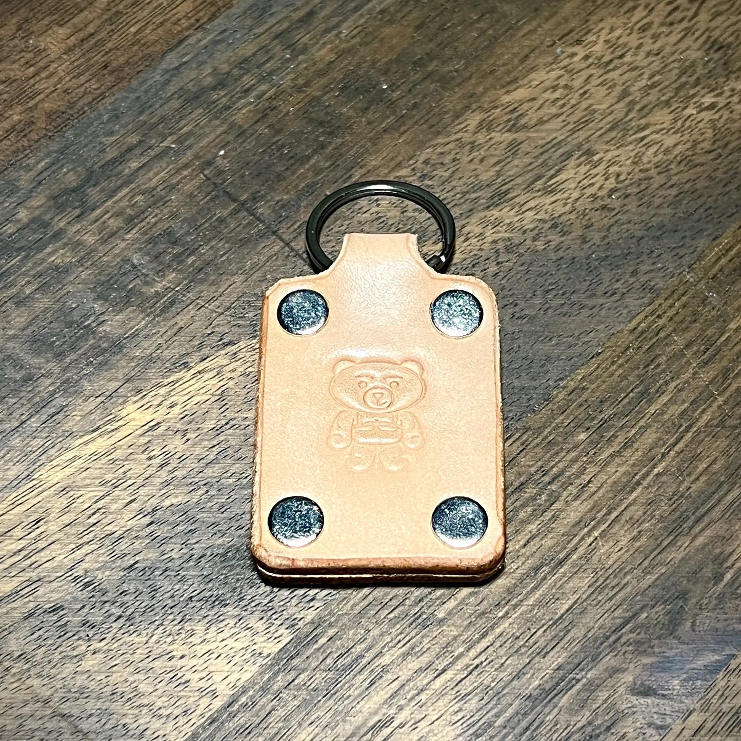 Front view of a natural leather keychain with four nickel-plated steel rivets, top split ring, and embossed bear logo.