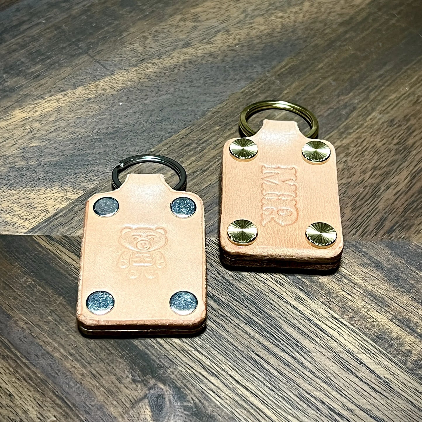 Hero tilt of natural leather keychains featuring embossed bear graphic and custom MIR initials with steel and brass hardware.