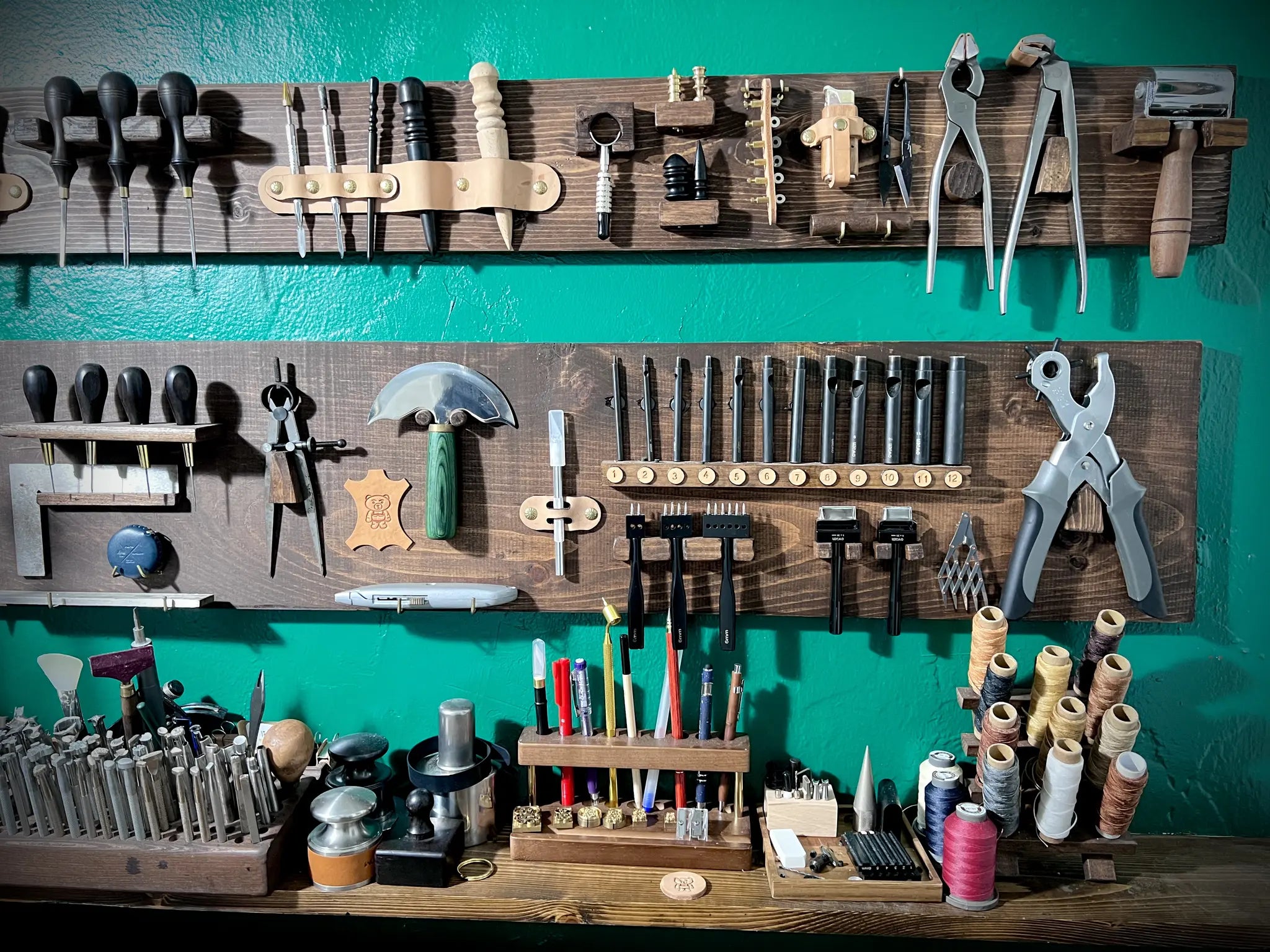 A detailed view of a leatherworking bench featuring an array of strap cutters, edge bevelers, punches, and heavy thread.