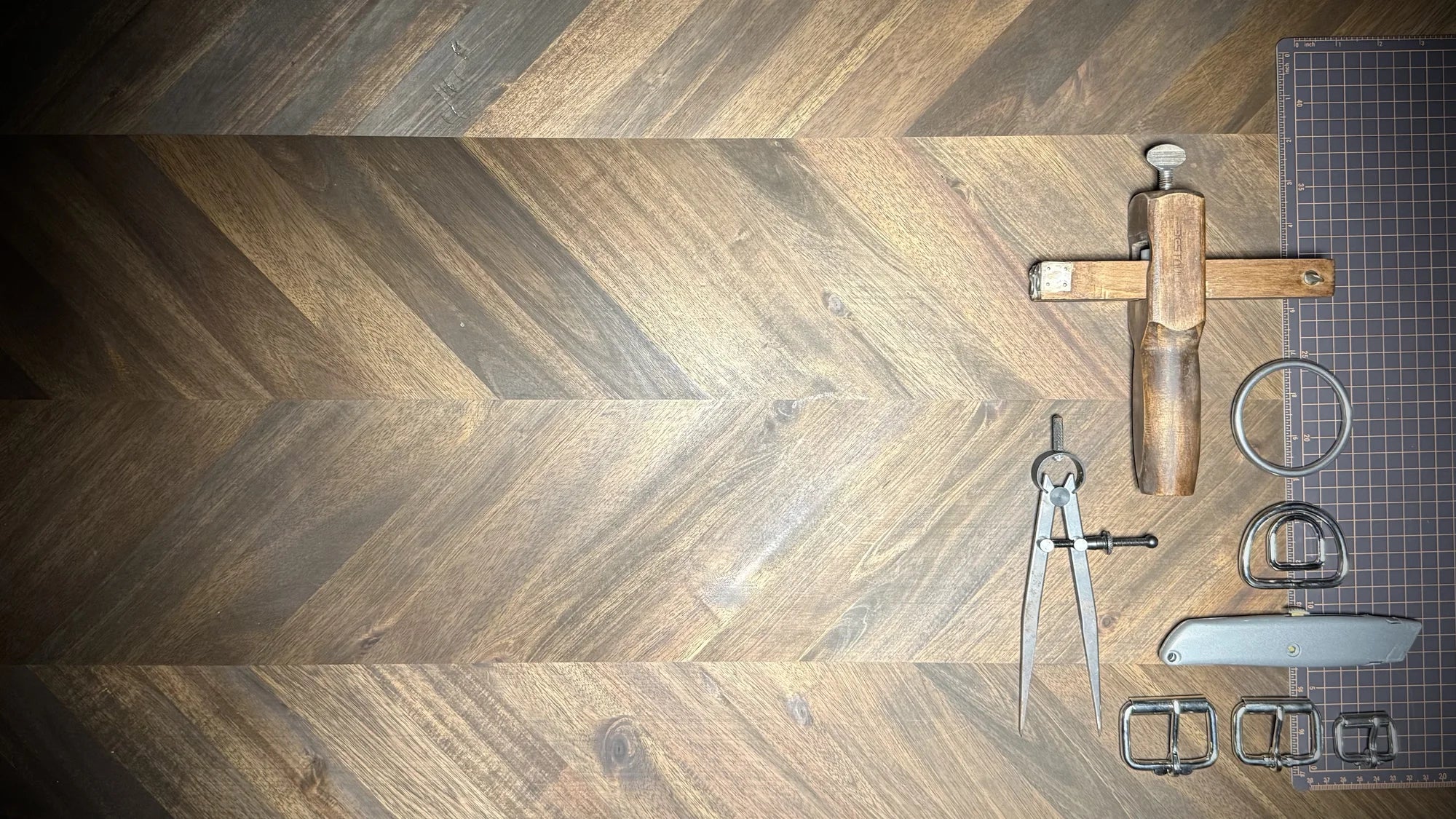 A collection of traditional leathercraft tools including a strap cutter, wing divider, and steel buckles laid out on a herringbone wood workbench.