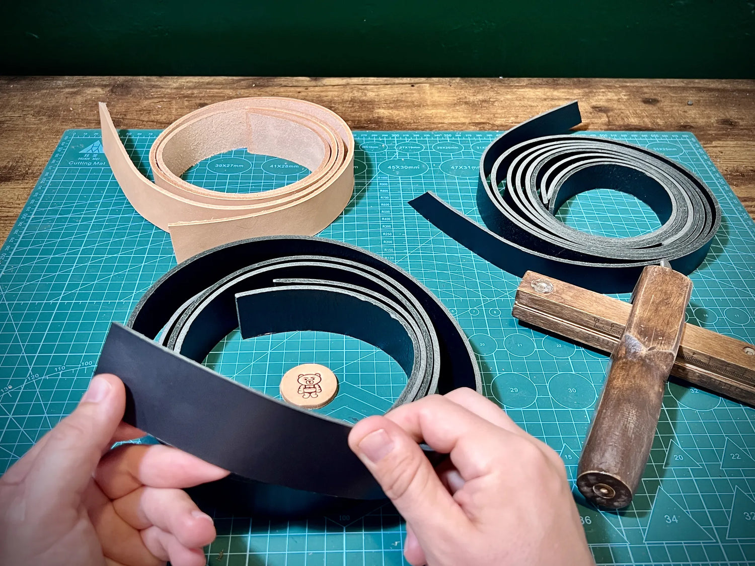Artisan hands measuring black and natural vegetable-tanned leather straps beside a wooden draw gauge on a green cutting mat.