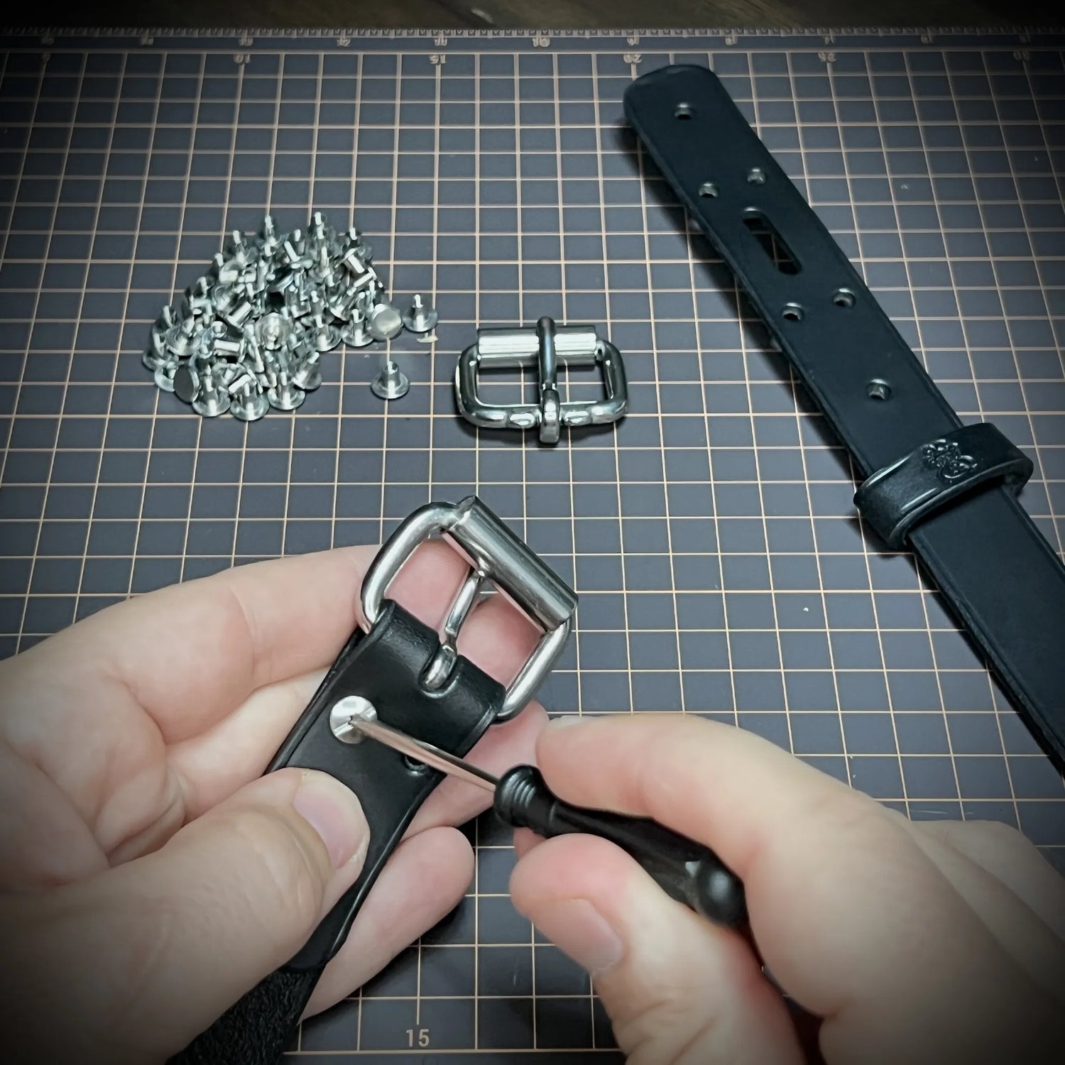 Close-up view of a maker manually securing a steel roller buckle onto a thick black leather cuff using a screwdriver and heavy-duty hardware rivets.