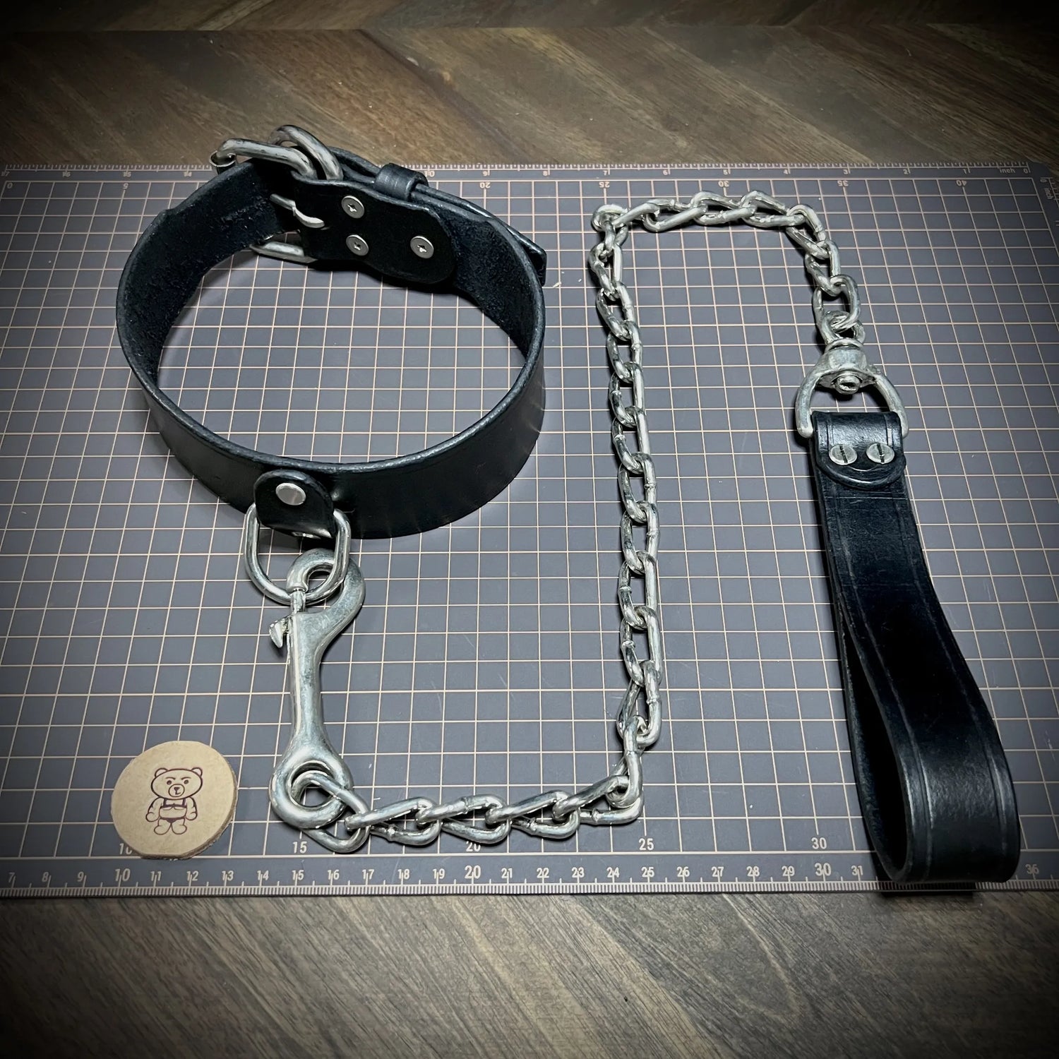 Full layout view of a completed 1.75 inches black leather posture collar connected to a heavy steel chain leash on a gridded workbench surface.