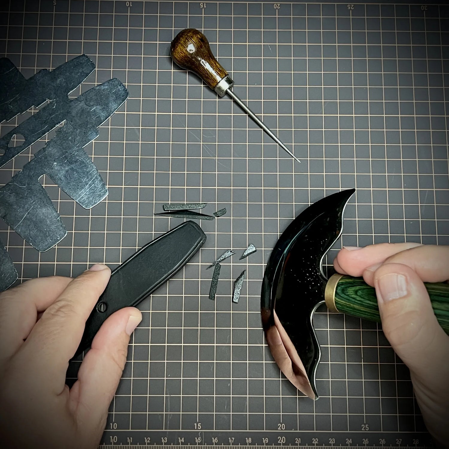 Close-up of a maker using a traditional steel half-moon knife to manually trim the edges of an 8oz black leather strap on a gridded studio mat.