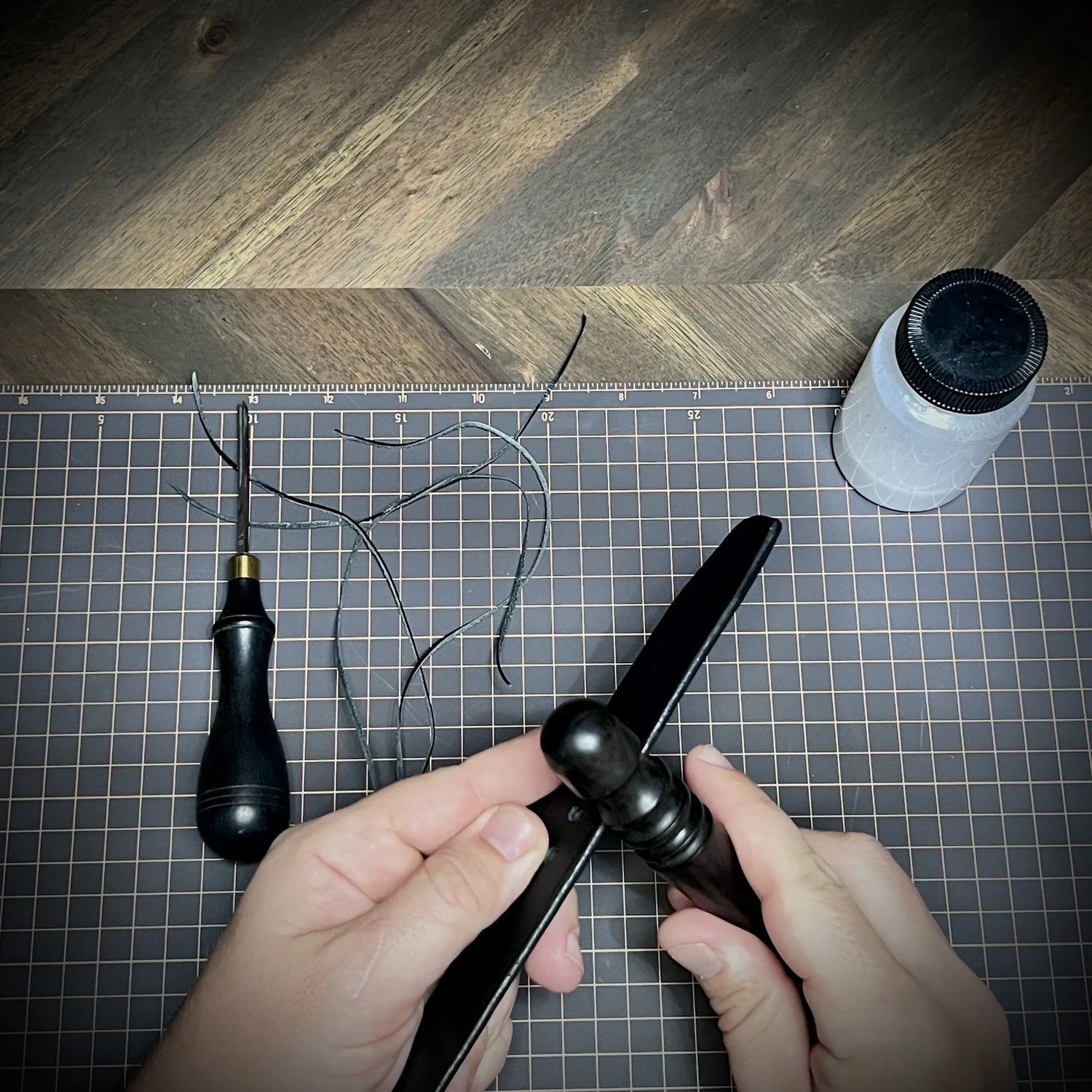 Close-up view of hands using a wooden edge slicker to burnish the edges of a heavyweight black leather strap with Tokonole on a workspace grid.