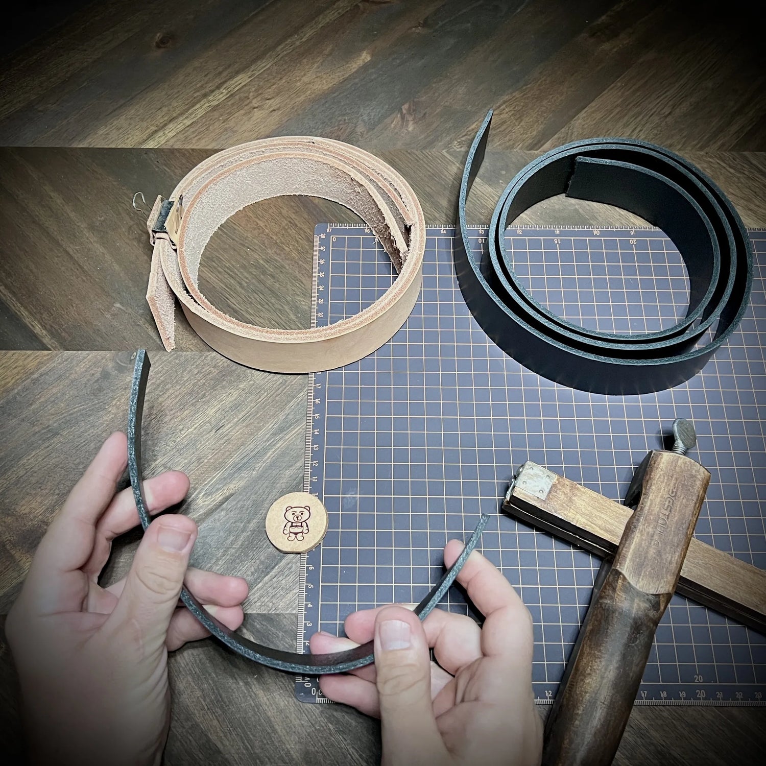 Top-down view of raw natural vegetable-tanned leather and dyed black leather straps on a gridded cutting mat during the sourcing and selection phase.