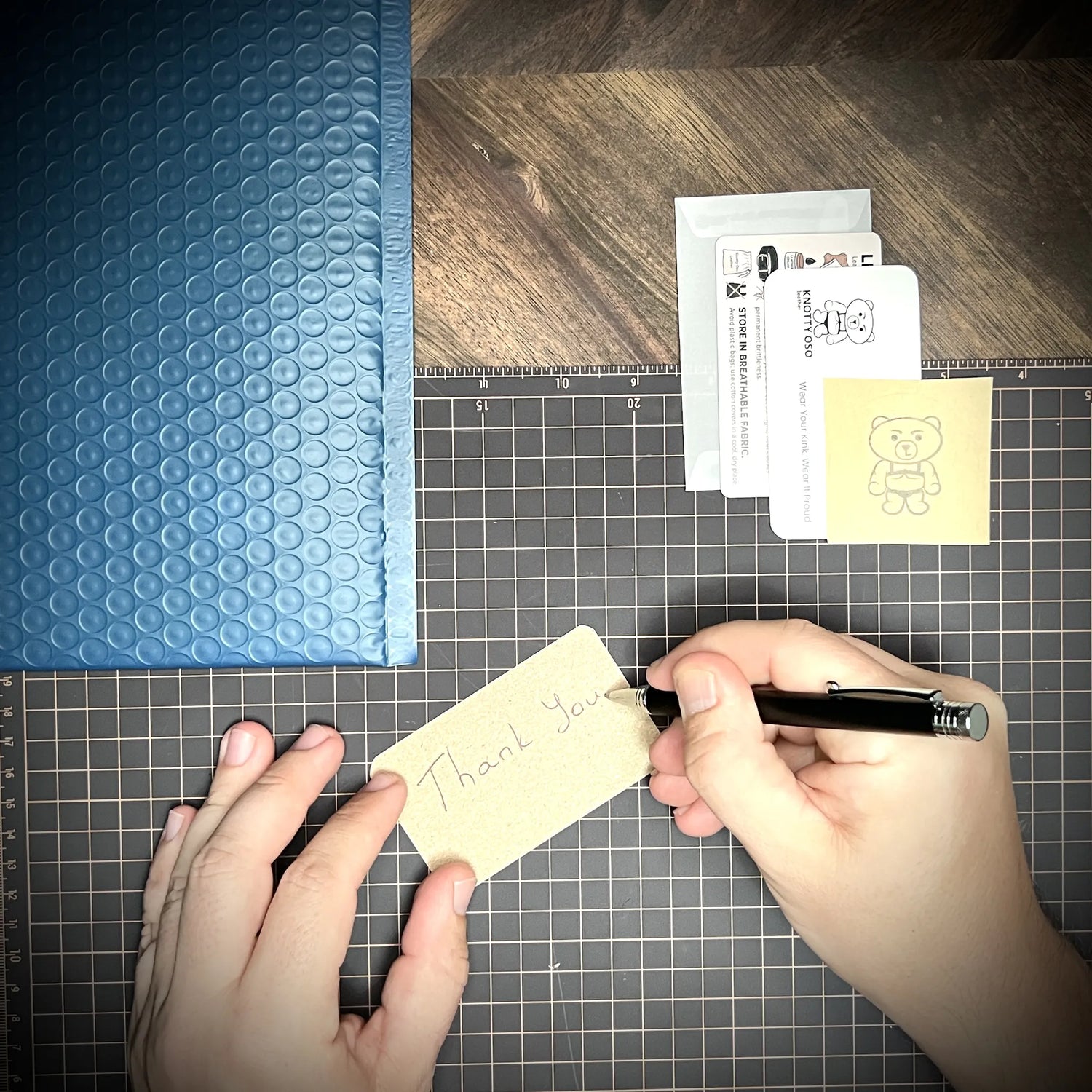 Top-down view of a maker writing a personalized note on a card next to a protective blue bubble mailer and branded studio cards on a cutting mat.