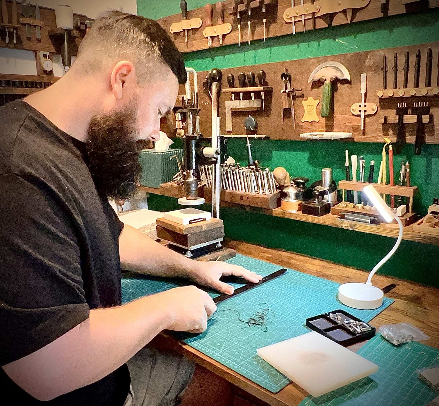 Artisan using a hand tool under a desk lamp to bevel edges on a custom strap at the wooden studio workbench.
