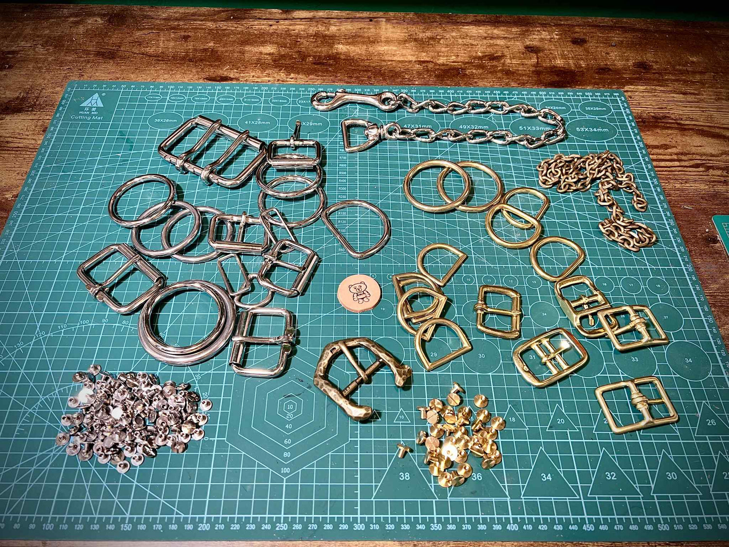 Array of solid brass buckles, steel D-rings, chains, snap hooks, and loose rivets arranged on a workshop mat.