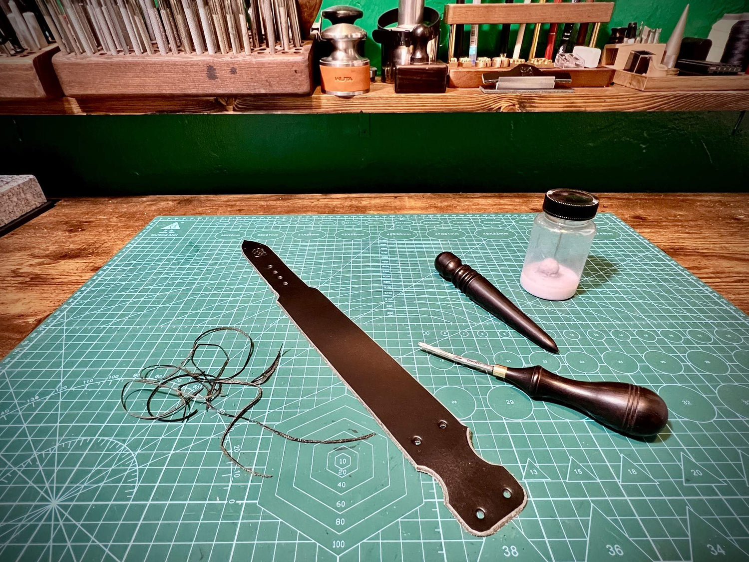 Black leather strap under construction on a cutting grid alongside edge slickers and professional beveling tools.