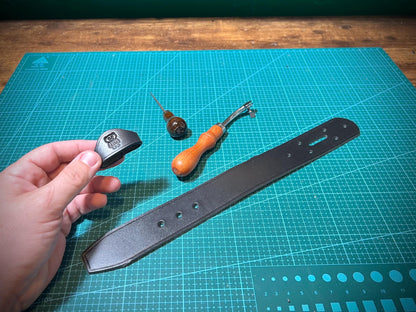 Hand holding a leather tool on a cutting mat with other tools and a strap.