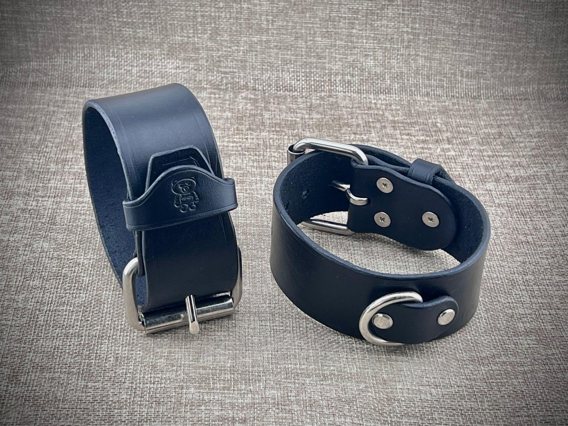 Two black leather cuffs with metal buckles on a textured gray background