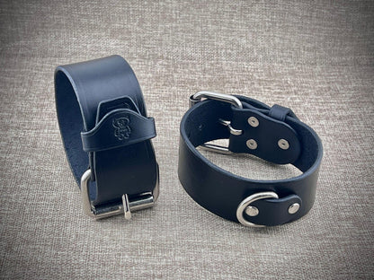 Two black leather cuffs with metal buckles on a textured gray background