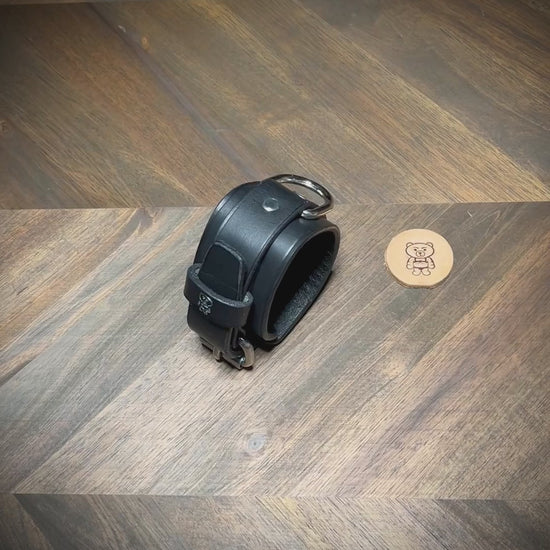 Overhead presentation of an artisan unbuckling a 1.75" black leather two-piece cuff with an integrated steel D-ring on a wood workbench surface.