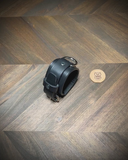 Overhead presentation of an artisan unbuckling a 1.75" black leather two-piece cuff with an integrated steel D-ring on a wood workbench surface.