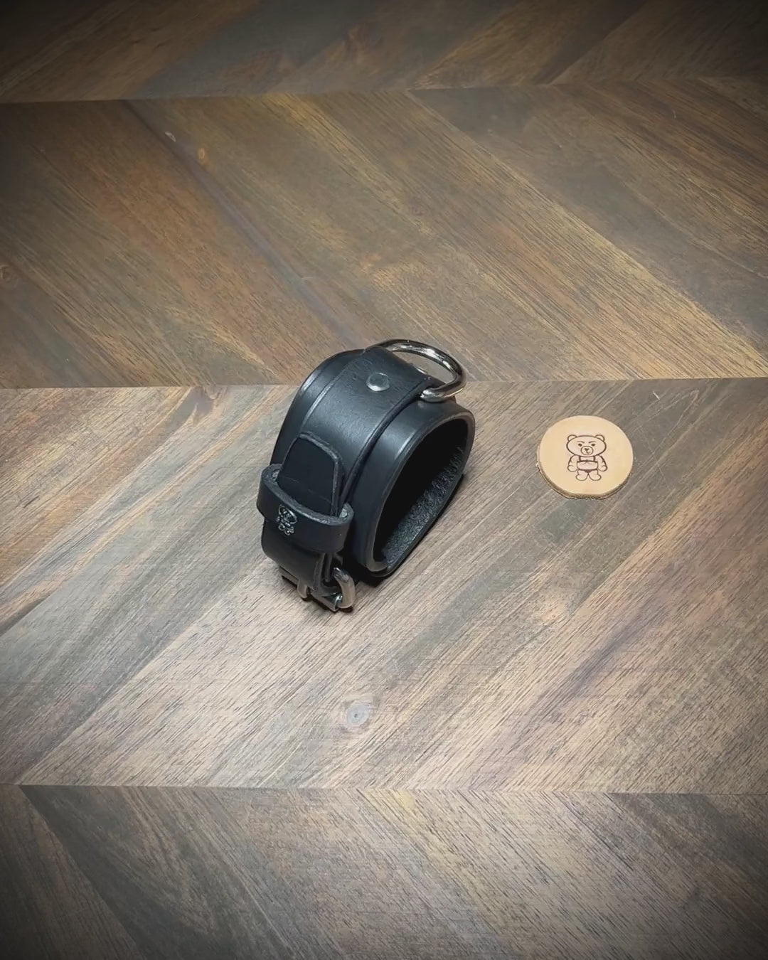 Overhead presentation of an artisan unbuckling a 1.75" black leather two-piece cuff with an integrated steel D-ring on a wood workbench surface.