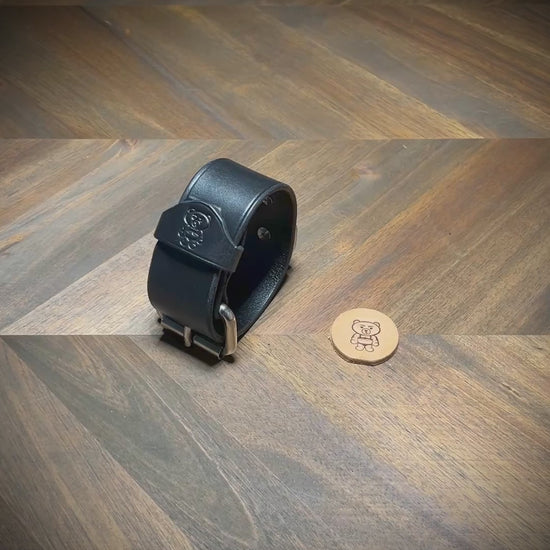 Hand-unbuckling and unrolling a 1.5" black leather cuff on a wood workbench to demonstrate hardware flexibility and interior grain texture.