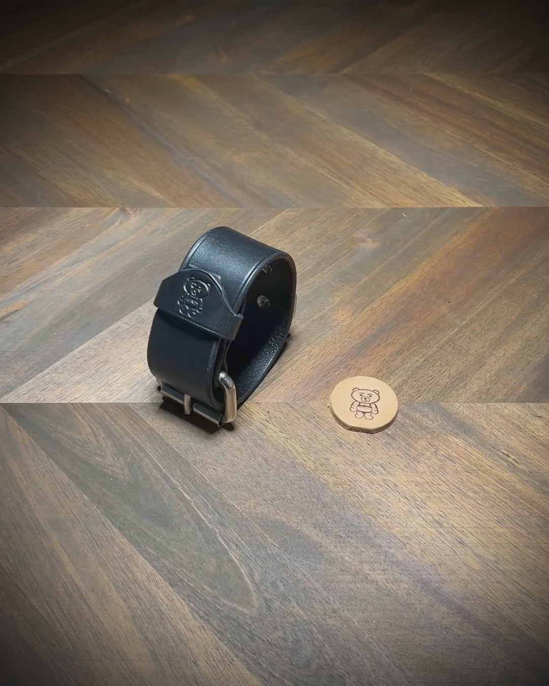 Hand-unbuckling and unrolling a 1.5" black leather cuff on a wood workbench to demonstrate hardware flexibility and interior grain texture.