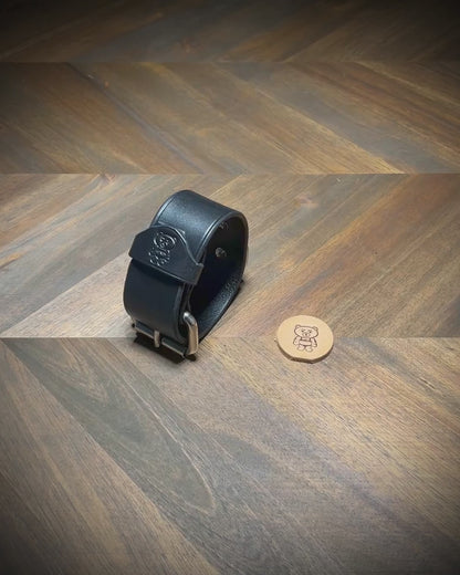Hand-unbuckling and unrolling a 1.5" black leather cuff on a wood workbench to demonstrate hardware flexibility and interior grain texture.