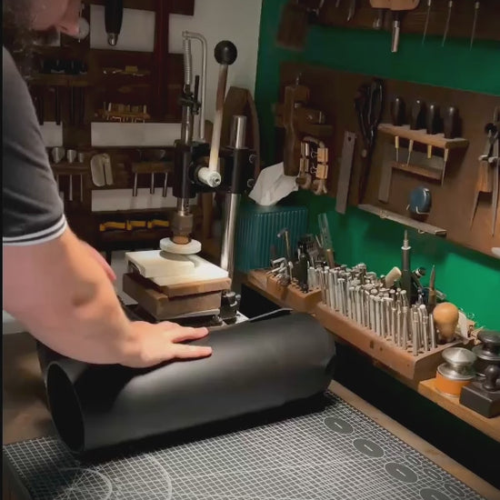 Bearded artisan at Knotty Oso, Miami Beach, hand-crafting premium black veg-tanned leather accessory with hand tools at a workshop bench.