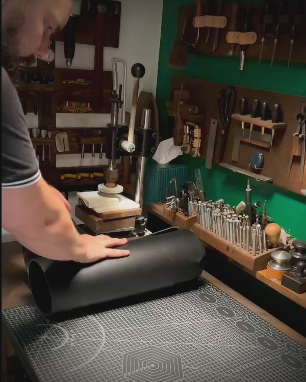 Bearded artisan at Knotty Oso, Miami Beach, hand-crafting premium black veg-tanned leather accessory with hand tools at a workshop bench.
