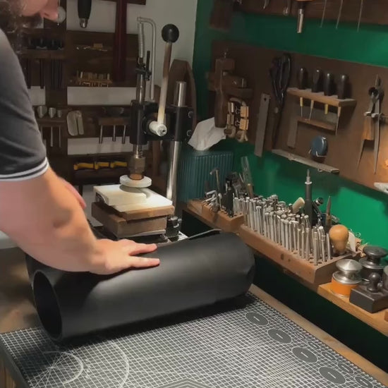Bearded artisan at Knotty Oso, Miami Beach, hand-crafting premium black veg-tanned leather accessory with hand tools at a workshop bench.