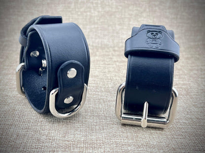 Two black leather cuffs with metal buckles on a textured gray background