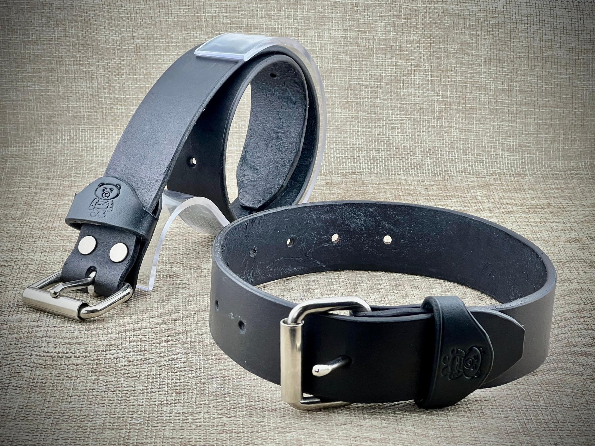 Two leather bondage restraints with metal buckles on a textured fabric background